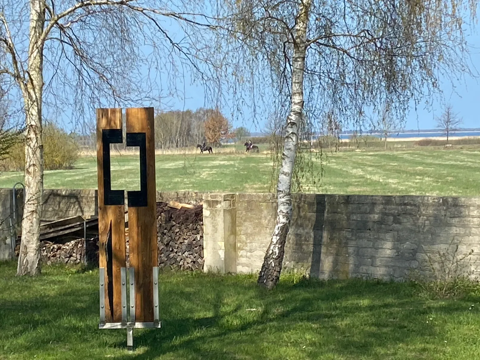 Holzskulptur im Garten, dahinter weite Wiesen und Birken
