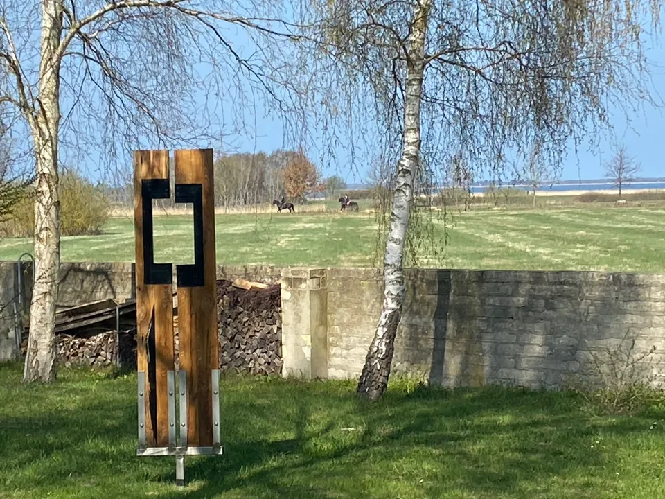 Holzskulptur im Garten, dahinter weite Wiesen und Birken